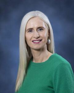 Professional woman with long blonde hair wearing a green top, smiling, in studio portrait.