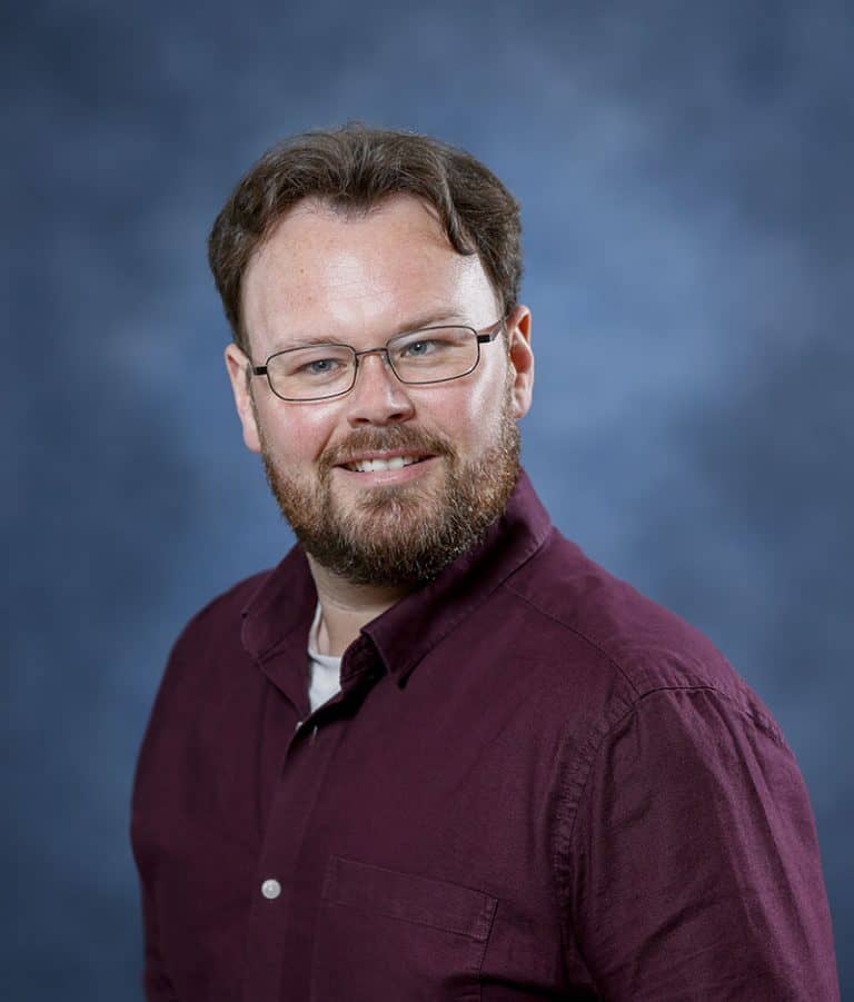A smiling man with glasses and a beard, wearing a maroon shirt, against a blue background.