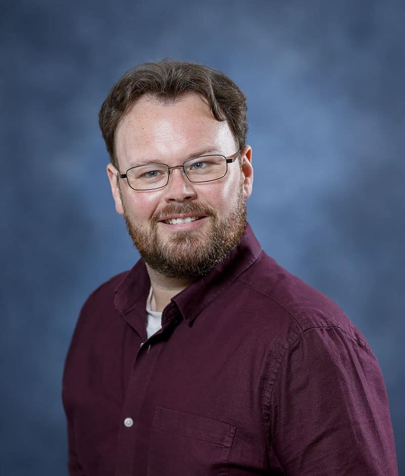 A smiling man with glasses and a beard, wearing a maroon shirt, against a blue background.