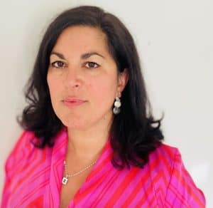 A woman with dark hair wearing a pink patterned top, standing against a plain background.