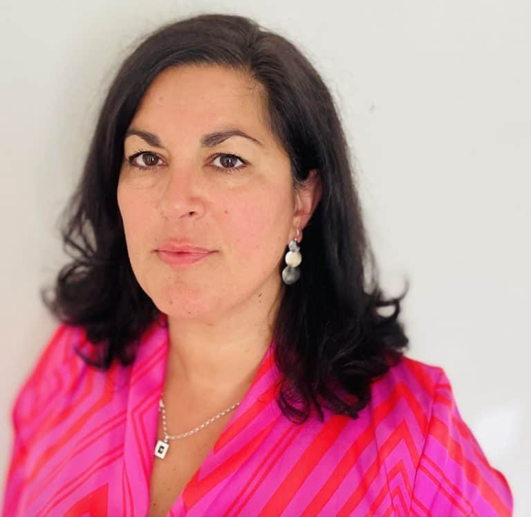A woman with dark hair wearing a pink patterned top, standing against a plain background.
