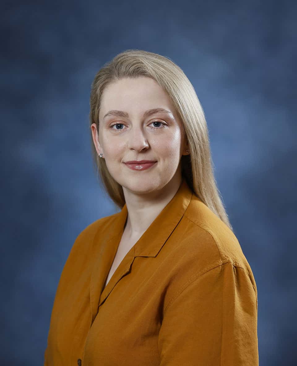 Professional woman with blonde hair in a mustard blouse against a blue background.