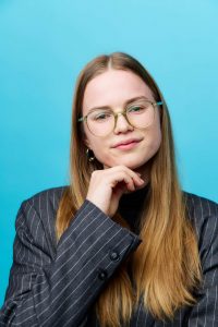 Thoughtful woman with glasses and long hair in a pinstripe blazer against blue background.