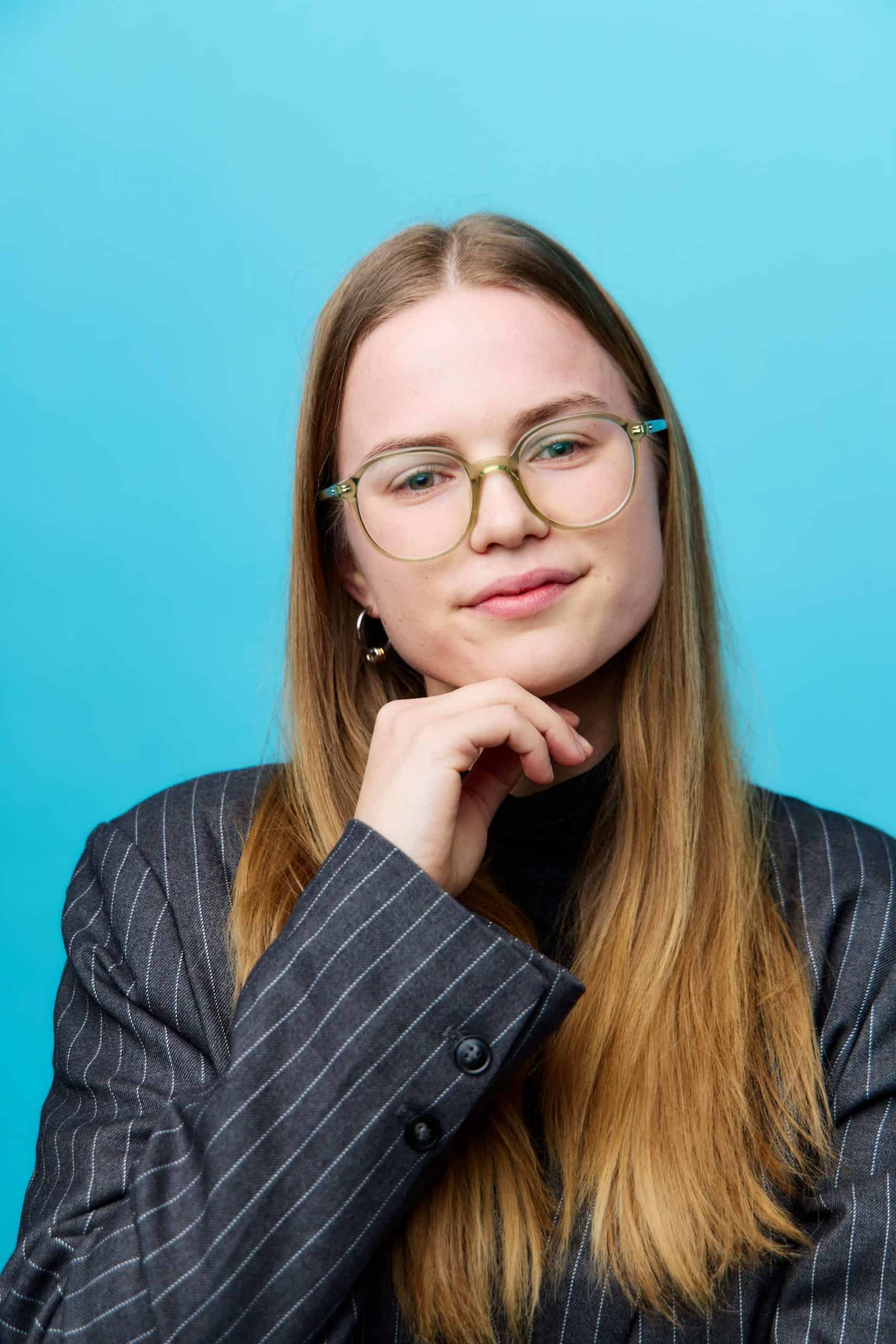 Thoughtful woman with glasses and long hair in a pinstripe blazer against blue background.