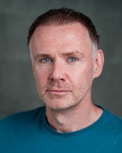 Close-up of a middle-aged man with short brown hair and blue eyes wearing a blue shirt.