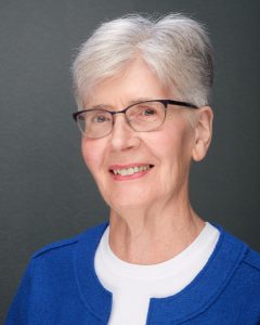 Smiling elderly woman with short grey hair and glasses in a blue top.
