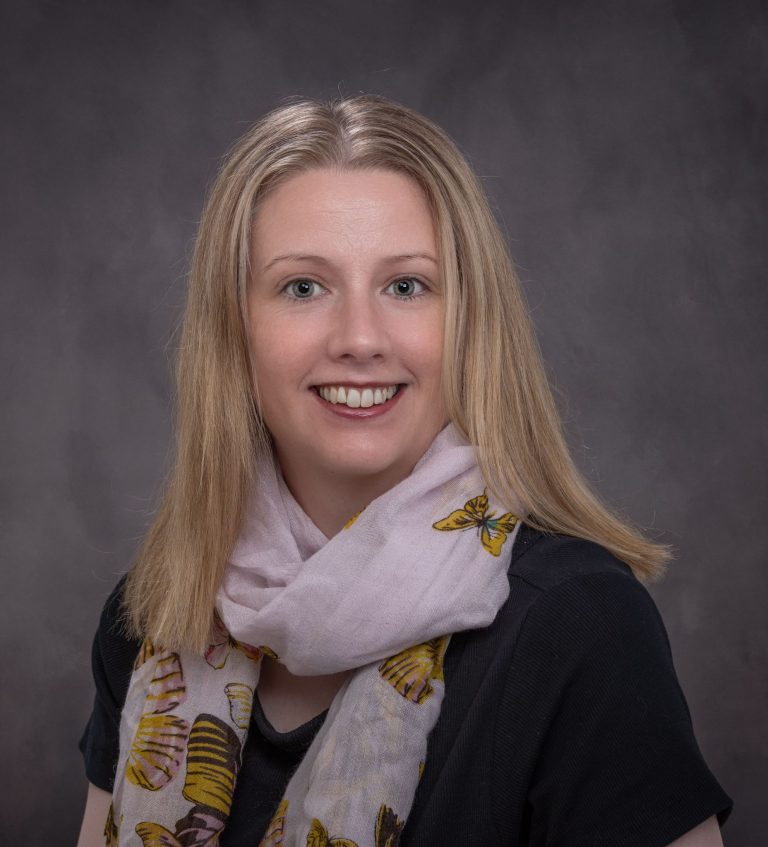 Smiling woman with blonde hair wearing a scarf and black top against grey background.