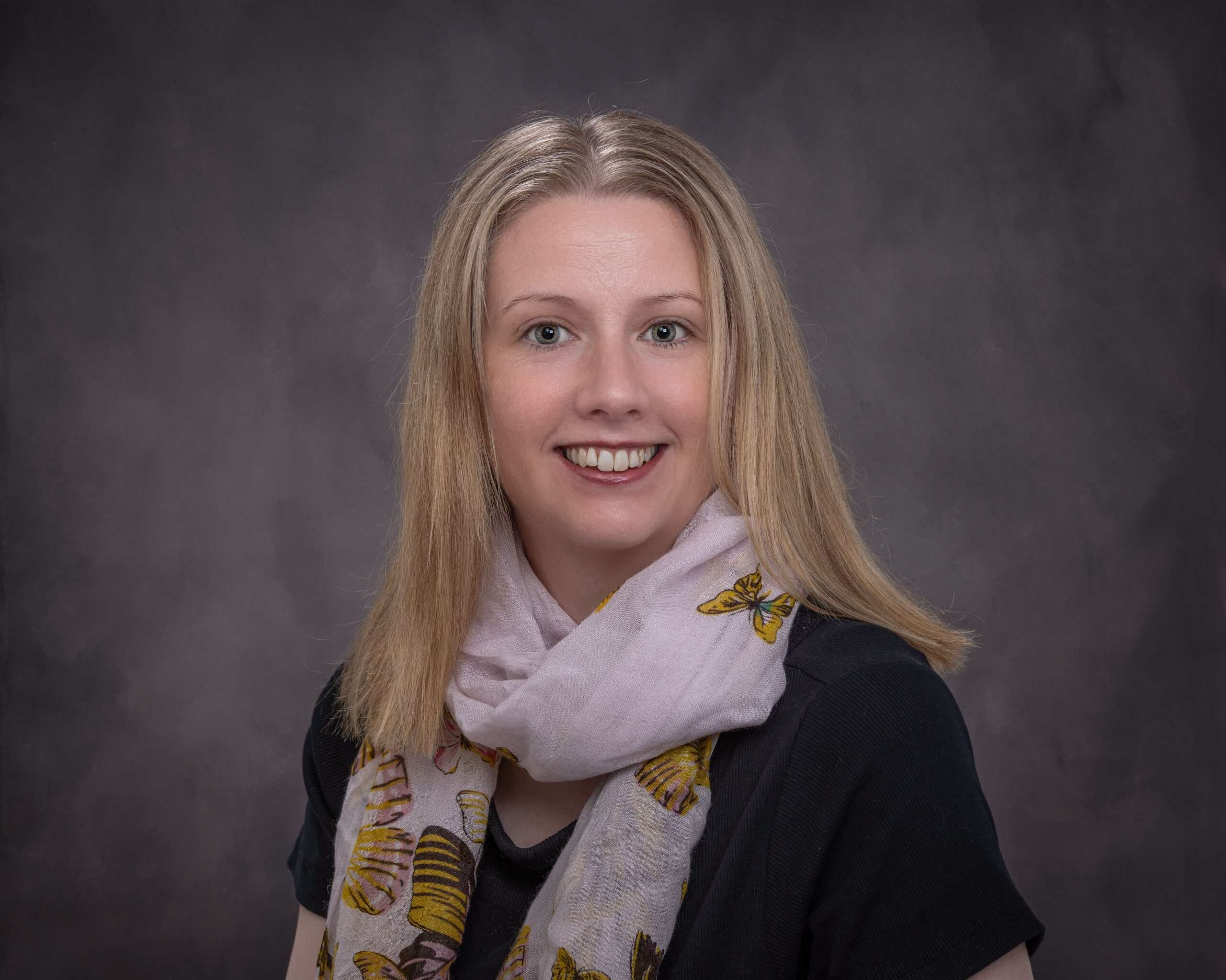Smiling woman with blonde hair wearing a scarf and black top against grey background.