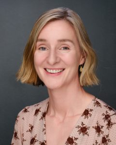 Smiling woman with shoulder-length blonde hair in a floral top against a dark background.