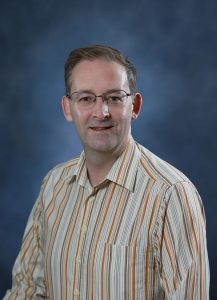 Professional portrait of a man in a striped shirt with glasses against a blue background.