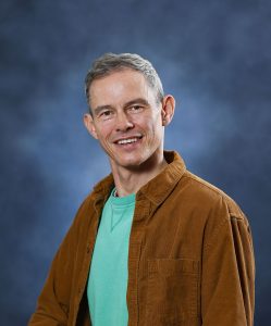 Smiling man in turquoise shirt and brown jacket against blue background.