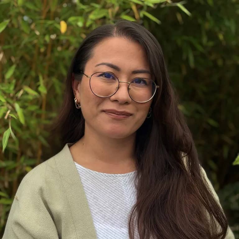 Professional woman with glasses and long dark hair outdoors in greenery.