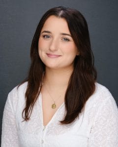 Young woman with long dark hair smiling in a white top against a grey background.