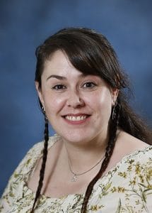 Smiling woman with dark hair and braids, wearing floral top, against blue background.
