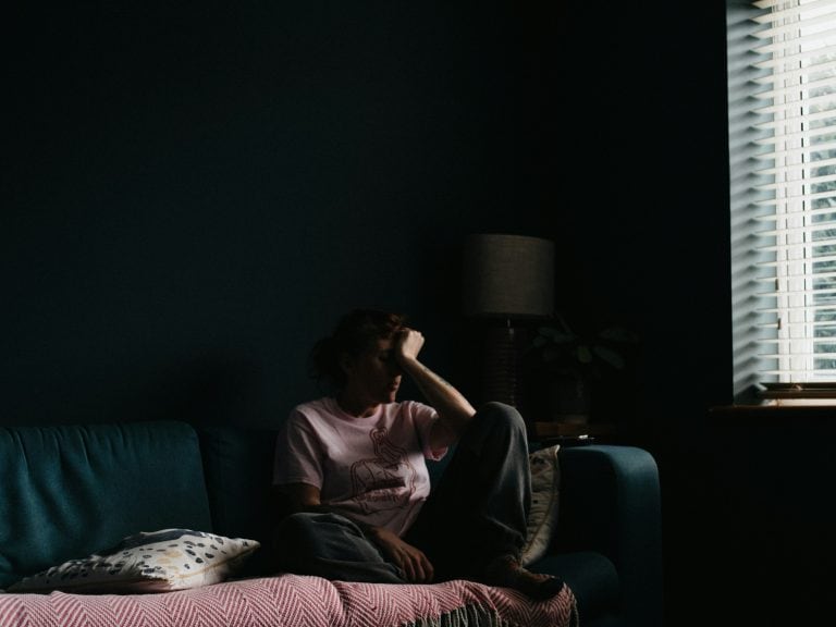 Woman sitting alone on a dark sofa in a dimly lit room, looking contemplative.